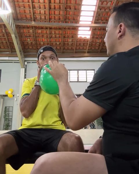 O aluno enchendo um balão, sentado, com o Nilberto ao lado.